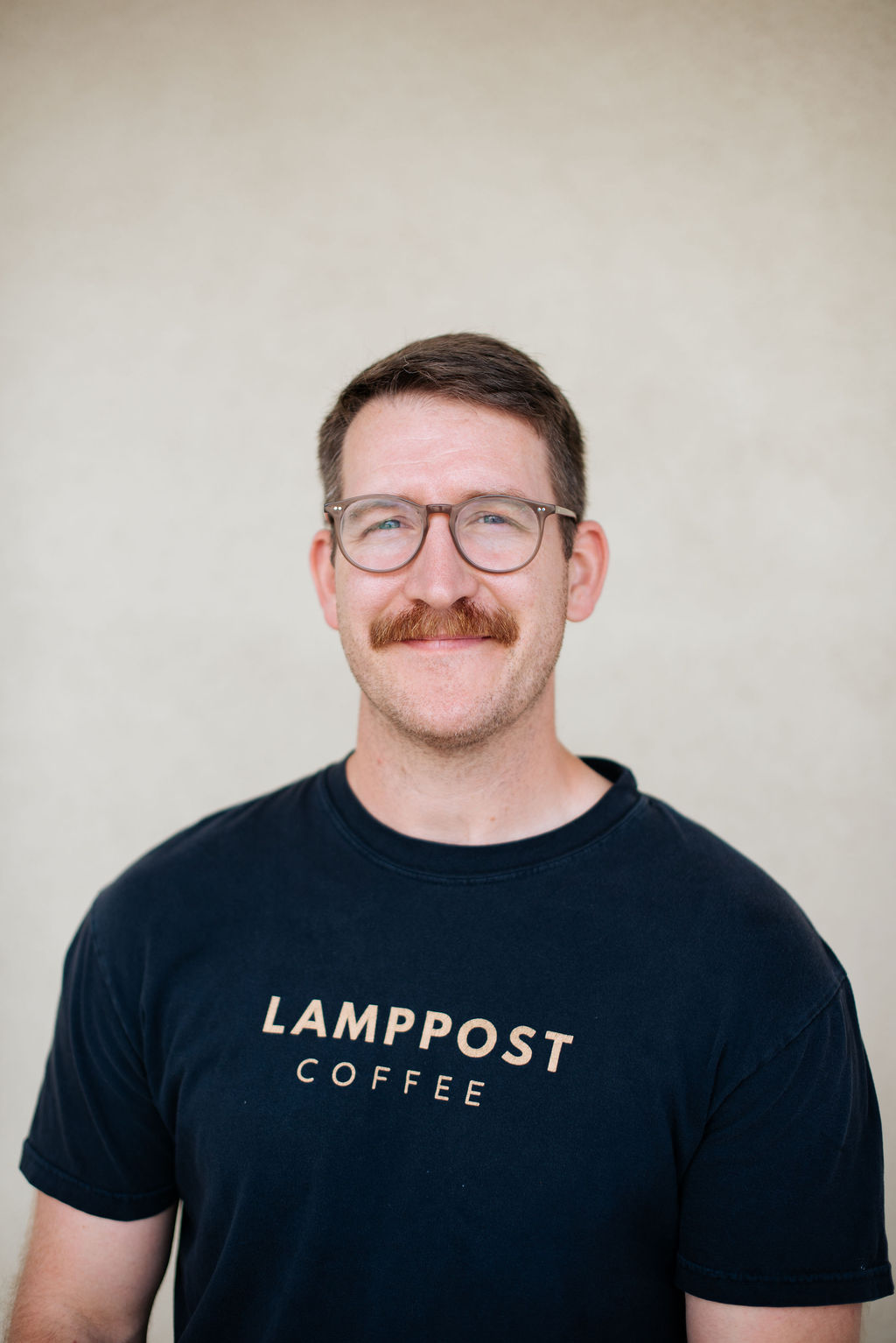 Aaron Anselmi, a man with short brown hair, glasses, and a mustache, wears a black "LAMPPOST COFFEE" T-shirt and stands against a plain light background. - Lamppost Coffee