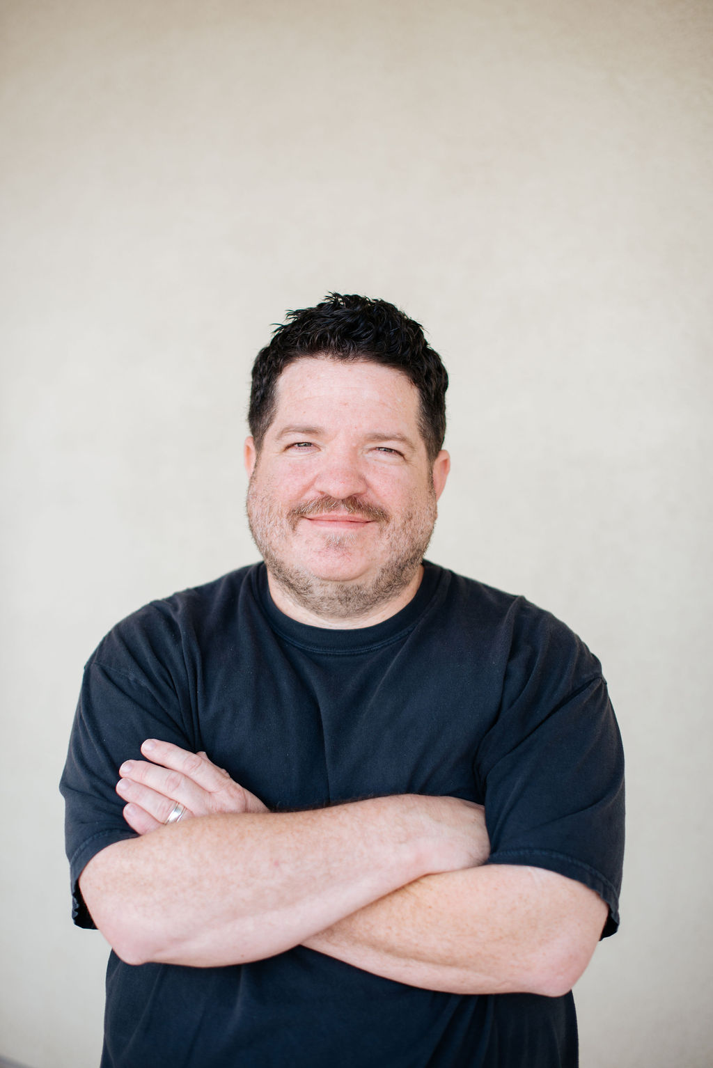 Man with short dark hair and a beard stands against a plain light-colored wall, wearing a black t-shirt and smiling with arms crossed. - Lamppost Coffee