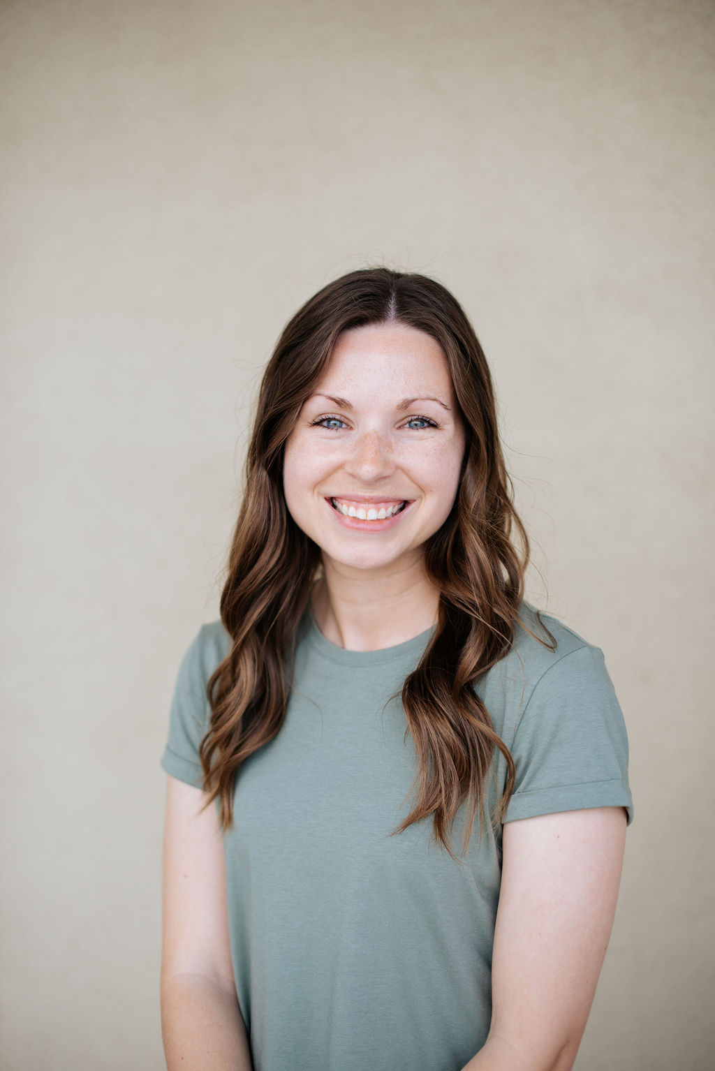 Mikella Hammonds, a woman with long brown hair and a green short-sleeve shirt, smiles at the camera in front of a plain beige background. - Lamppost Coffee