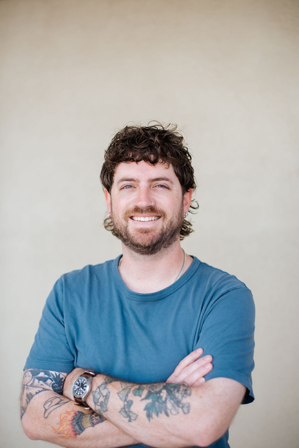 Pierce Hardin, a man with curly hair and a beard, stands against a plain background, smiling with arms crossed. He wears a blue t-shirt and displays tattoos on both arms. - Lamppost Coffee