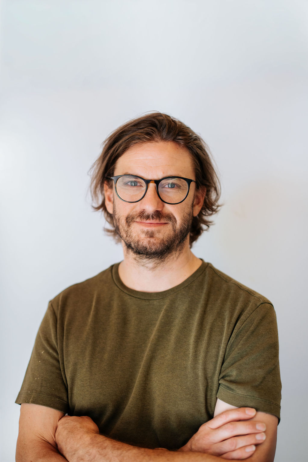 A man with brown hair and glasses, wearing an olive green t-shirt, stands with his arms crossed in front of a plain light background. - Lamppost Coffee