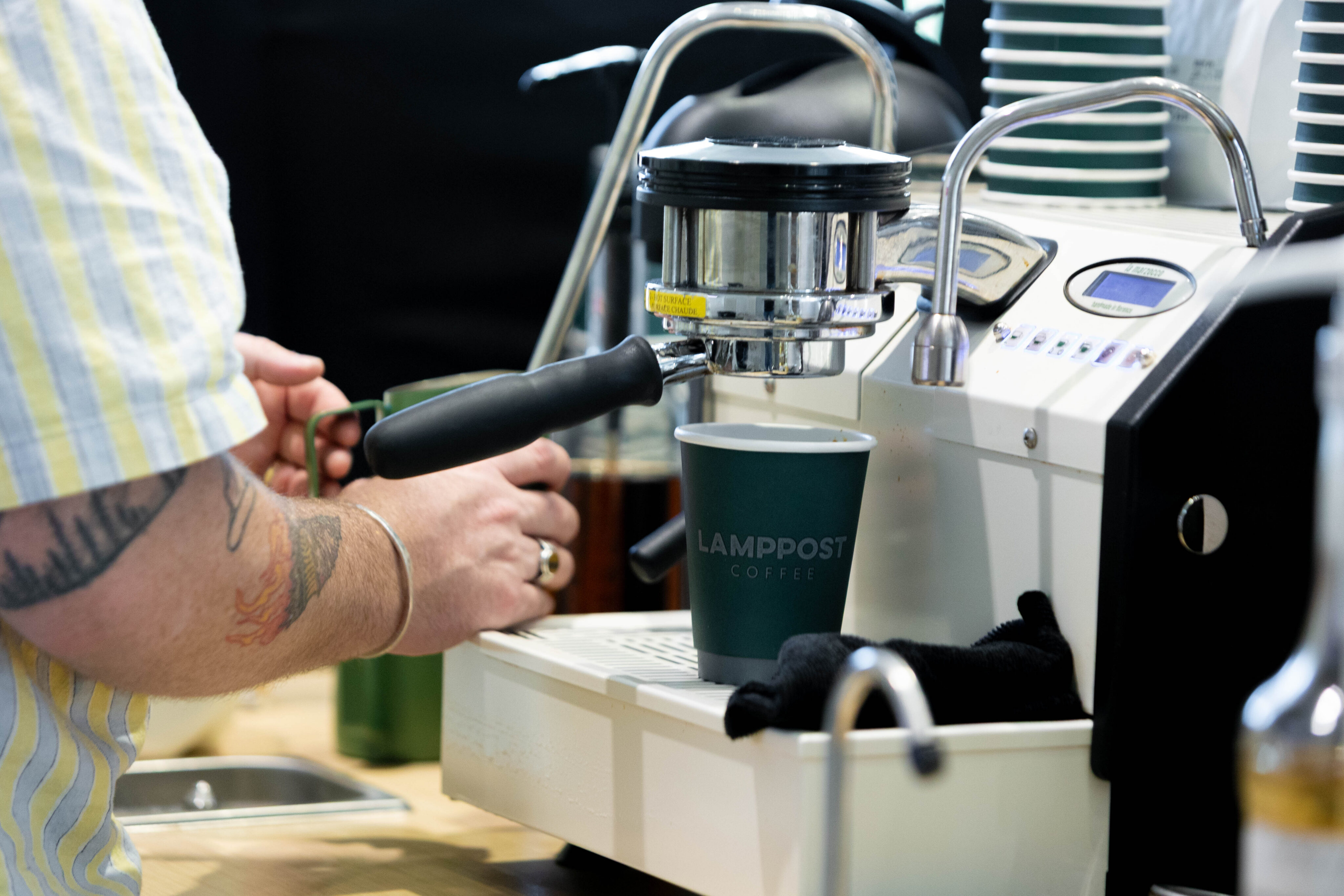 A person prepares coffee using an espresso machine, holding a cup labeled "Lampost Coffee" under the portafilter. - Lamppost Coffee