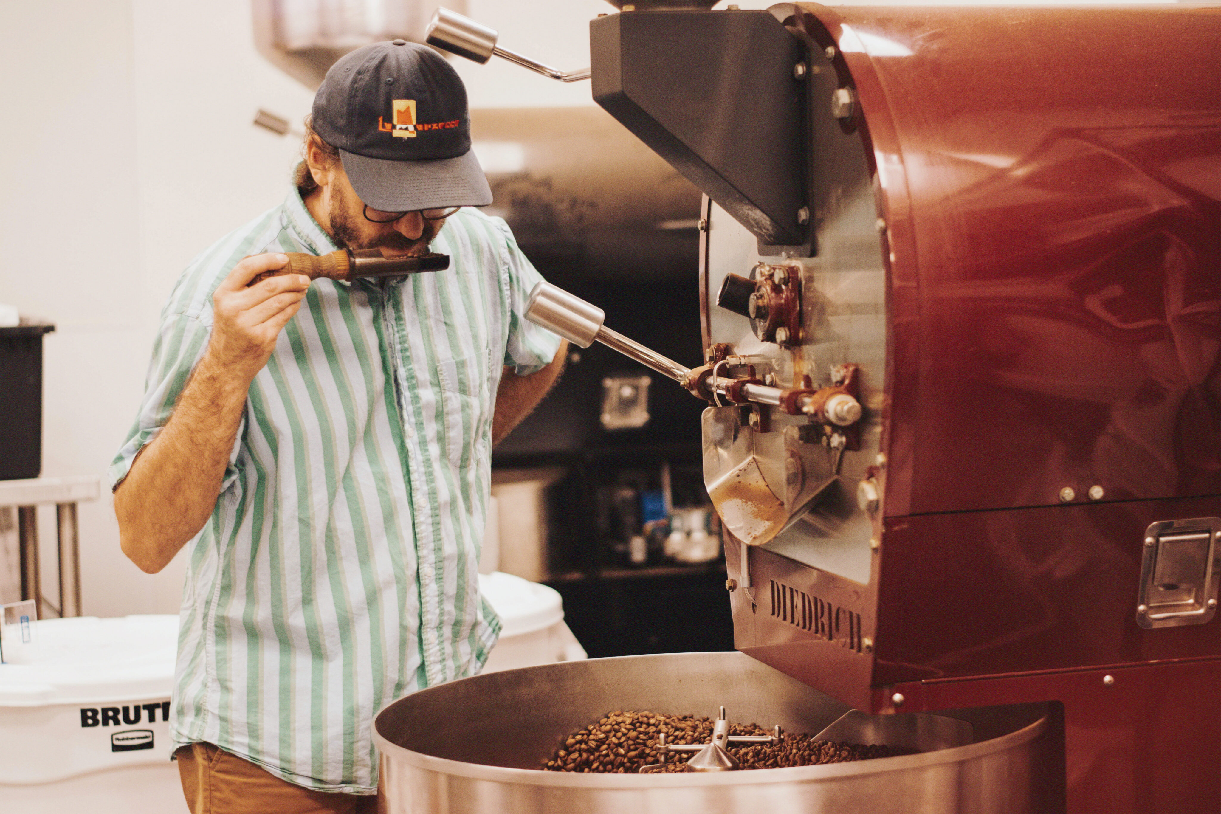 A man in a striped shirt and cap inspects roasted coffee beans by smelling them next to a large industrial coffee roasting machine inside a bustling roasting facility. - Lamppost Coffee