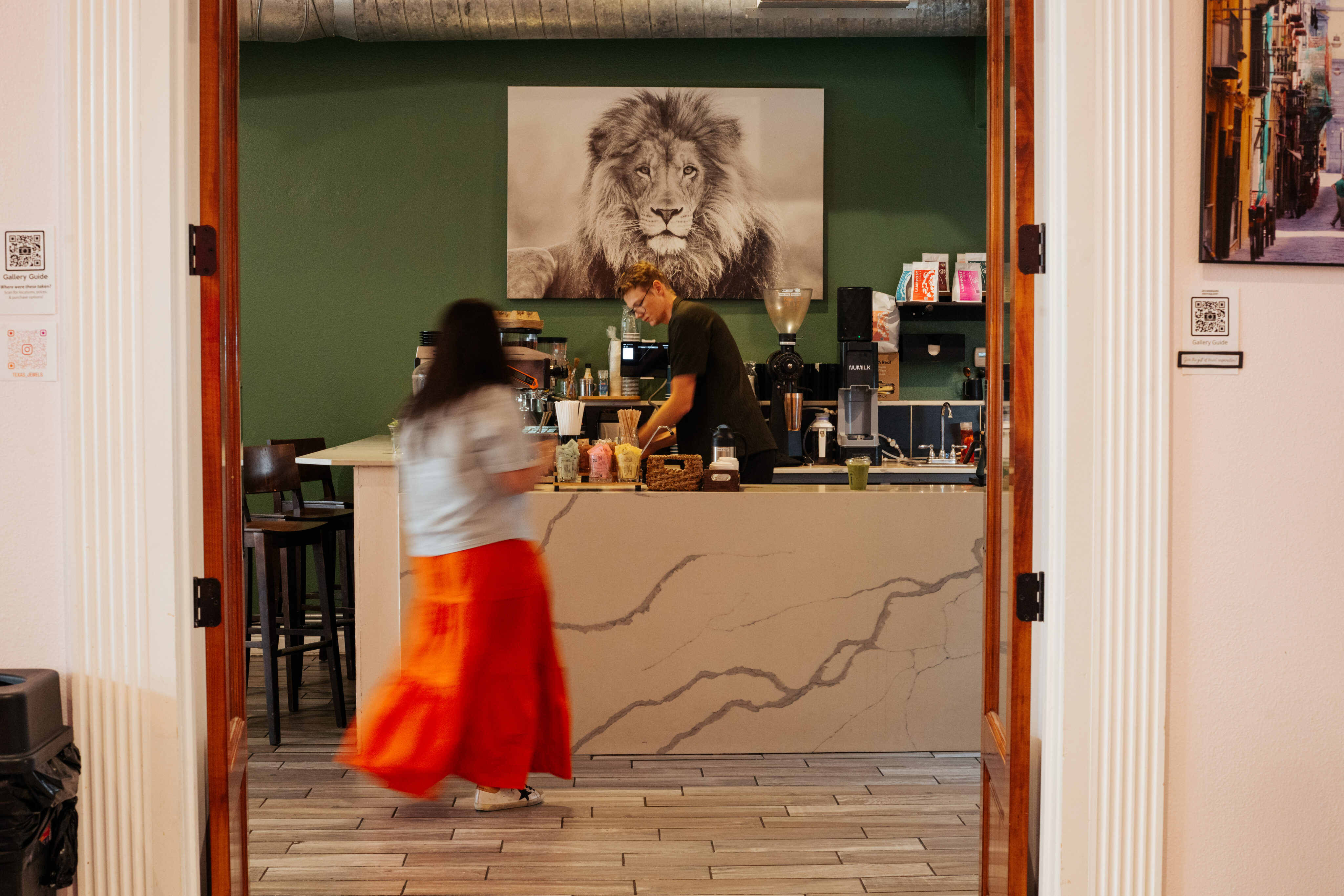 A woman in a red skirt approaches a cafe counter where a barista stands, with a large lion photo displayed on the green wall behind them. - Lamppost Coffee