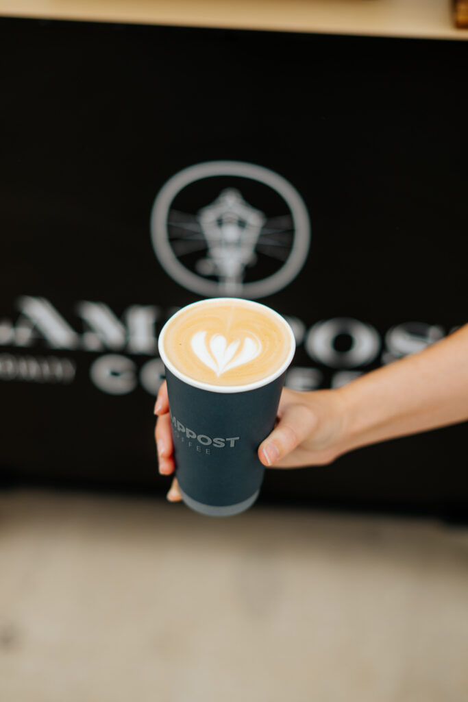 A hand holds a mobile coffee takeaway cup with heart-shaped latte art on top; a blurred logo and text appear in the background. - Lamppost Coffee
