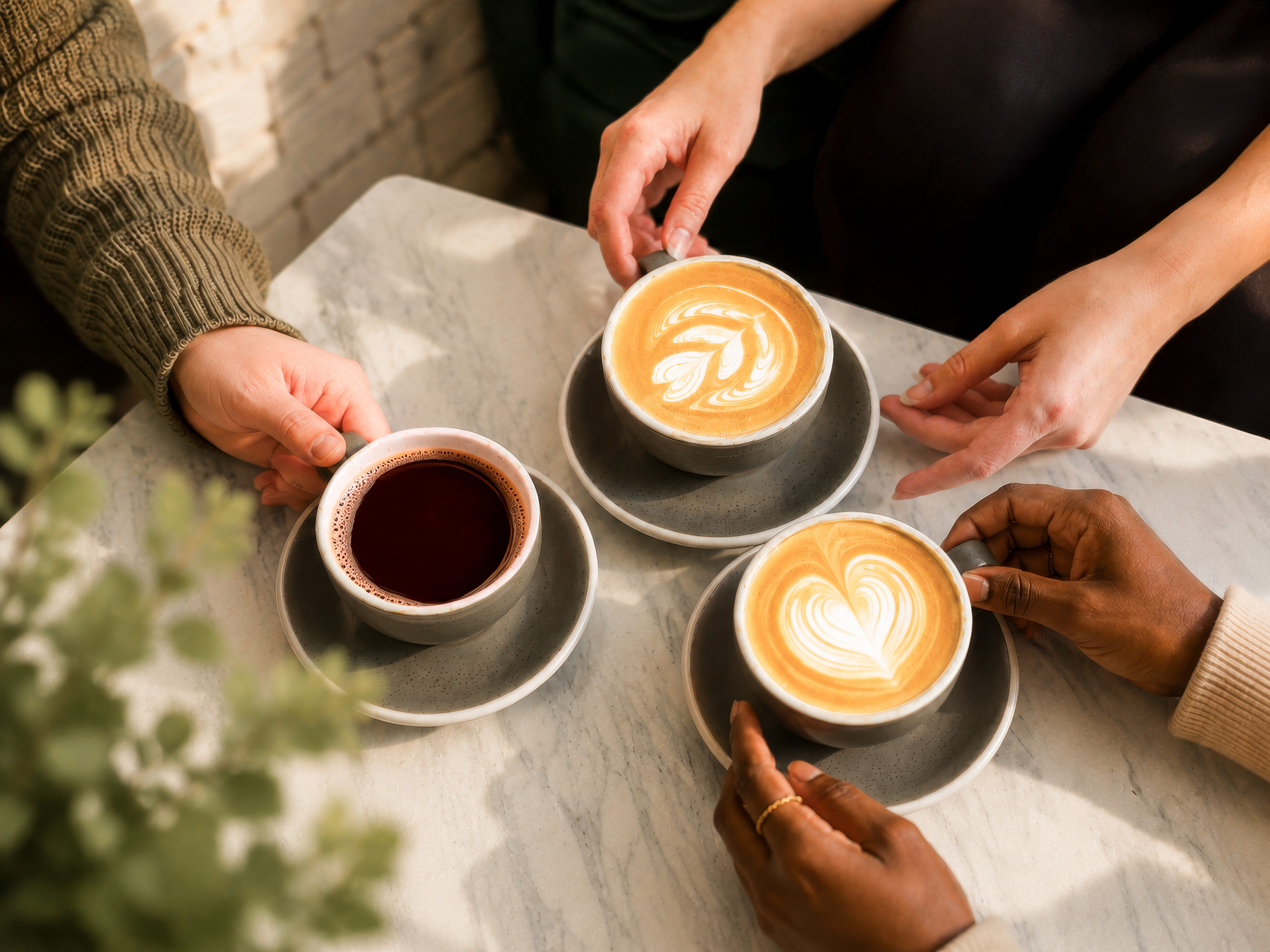 Three people enjoy Lamppost Coffee, holding cups of latte art and black coffee on a marble table. - Lamppost Coffee
