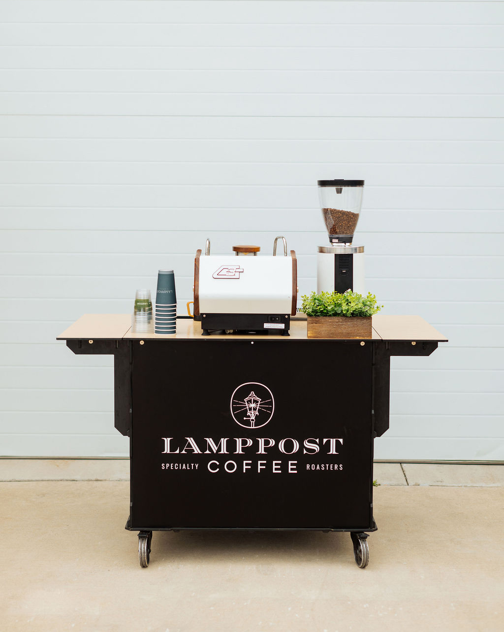 A mobile coffee cart labeled "Lamppost Coffee" features a compact espresso machine, cups, a grinder, and a potted plant—all set up for convenient mobile coffee service in front of a light-colored wall. - Lamppost Coffee