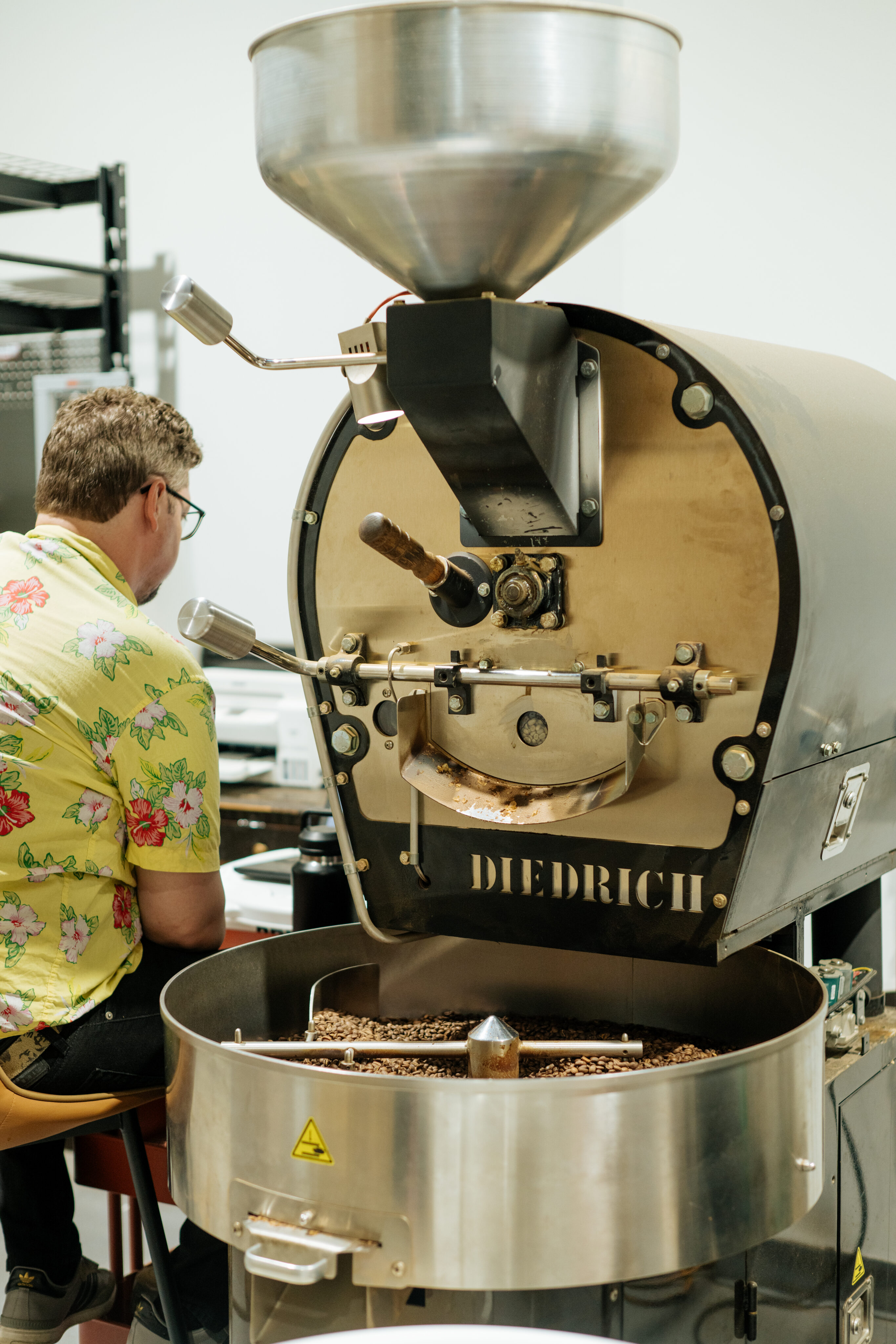 A man in a floral shirt sits beside a large Diedrich coffee roasting machine filled with roasted coffee beans in a workspace. - Lamppost Coffee
