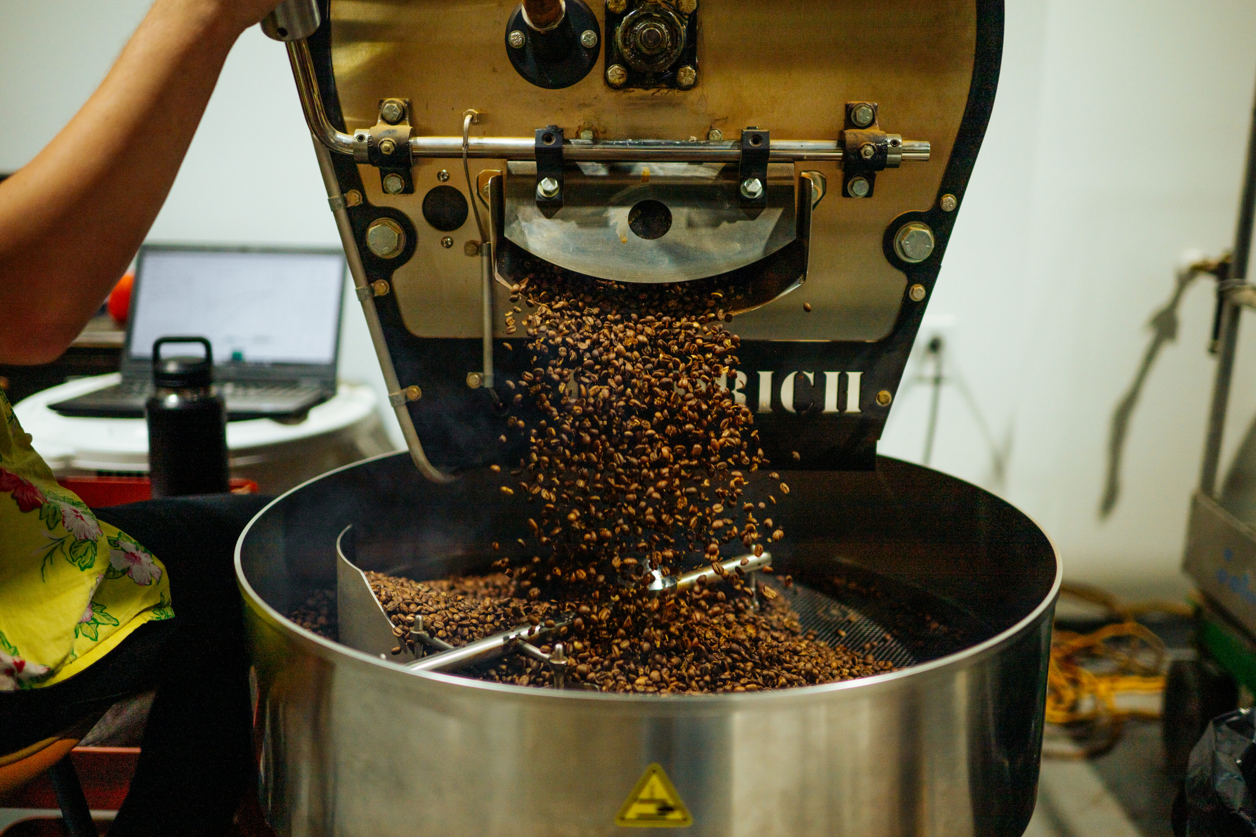 Freshly roasted coffee beans from Lamppost Coffee's commercial roasting machine tumble into a cooling tray, with a team member’s arm visible on the left. - Lamppost Coffee