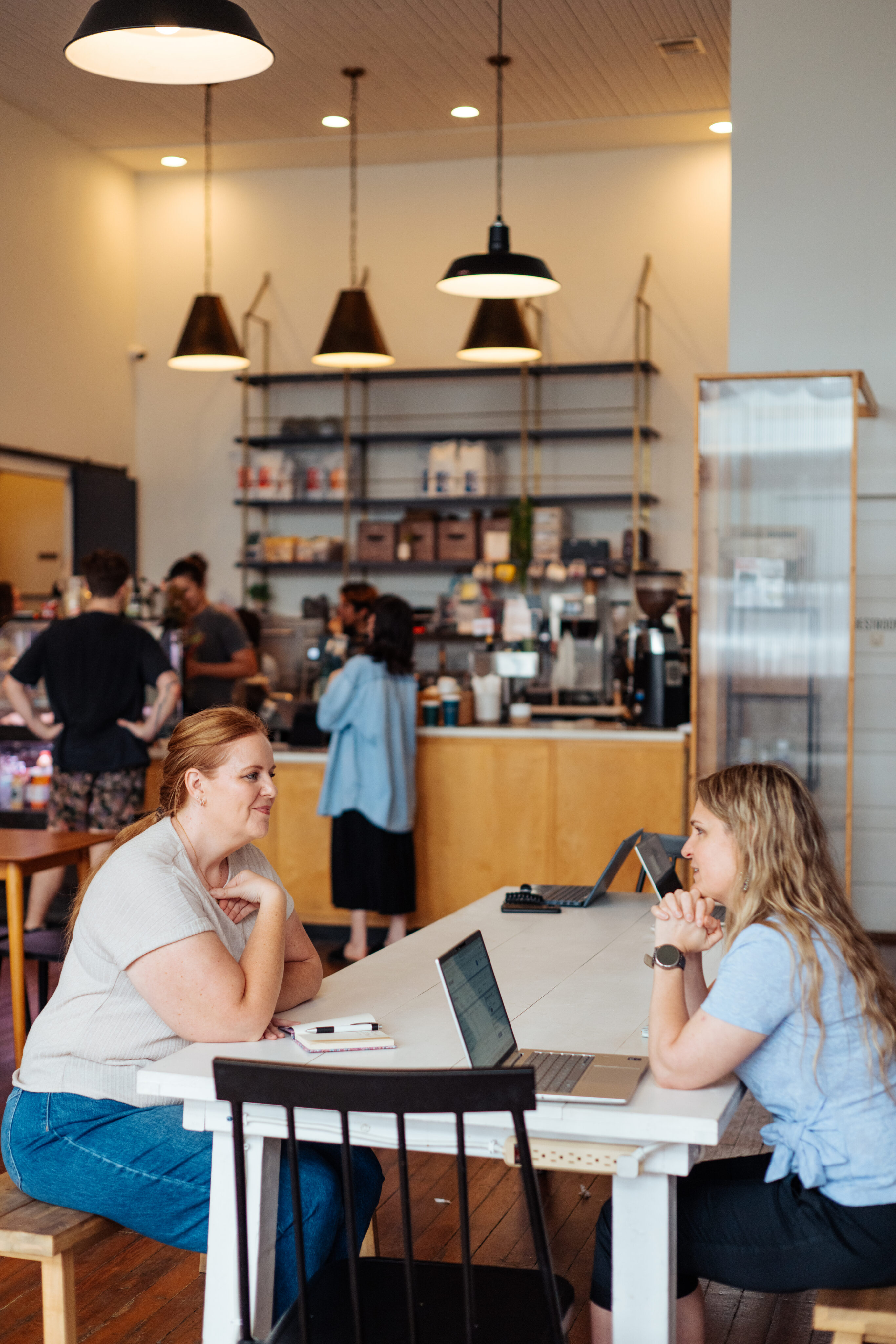 Two women sit across from each other at a white table with laptops in a coffee shop, while other customers and baristas are visible in the background. - Lamppost Coffee