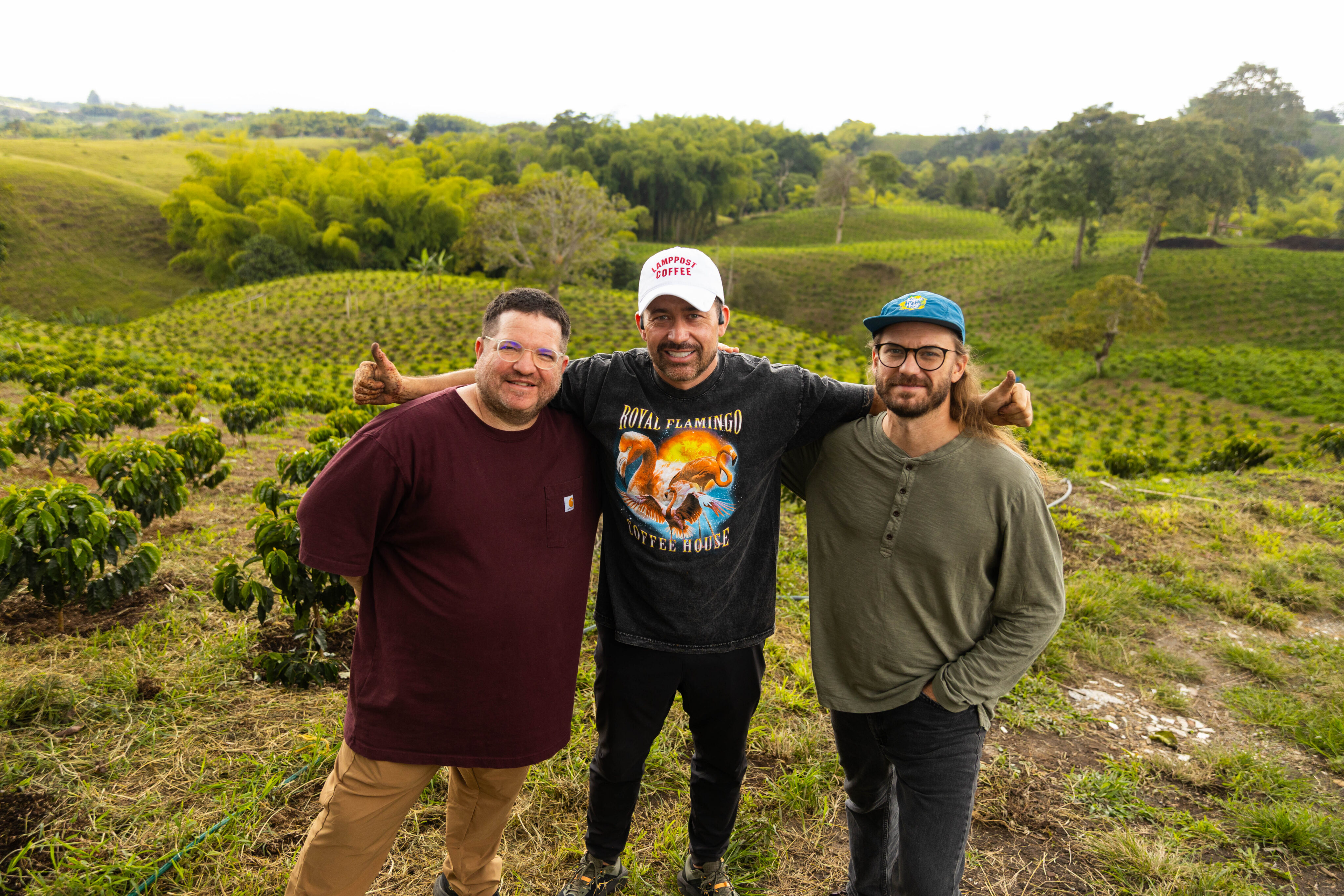 Three men, including Edwin Noreña, stand together smiling in front of a lush, green agricultural field with trees and hills in Colombia in the background. - Lamppost Coffee
