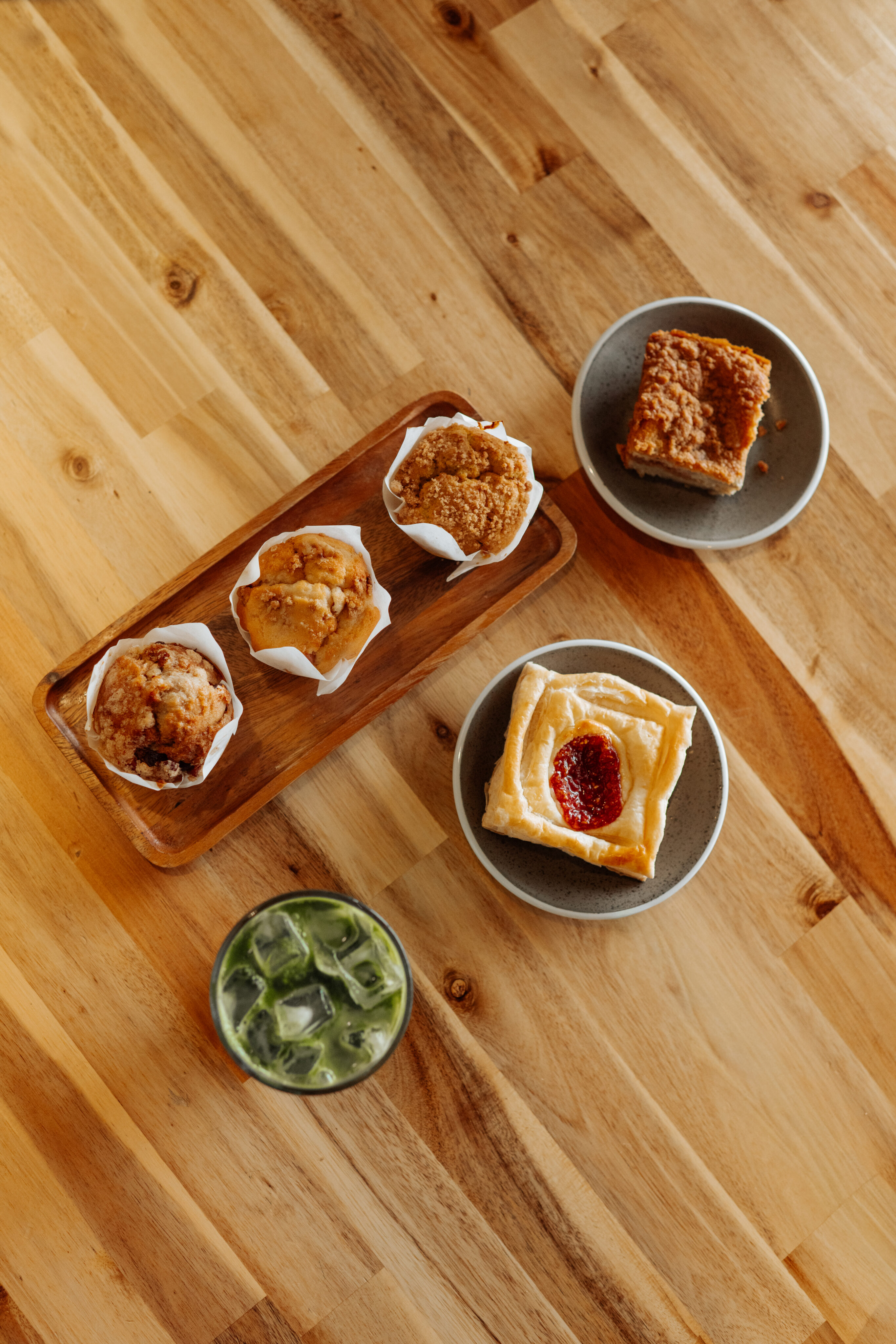 A wooden table set for catering features three muffins on a tray, a slice of coffee cake, a pastry with jam on a plate, and a glass of iced green drink—perfect for any food lover. - Lamppost Coffee
