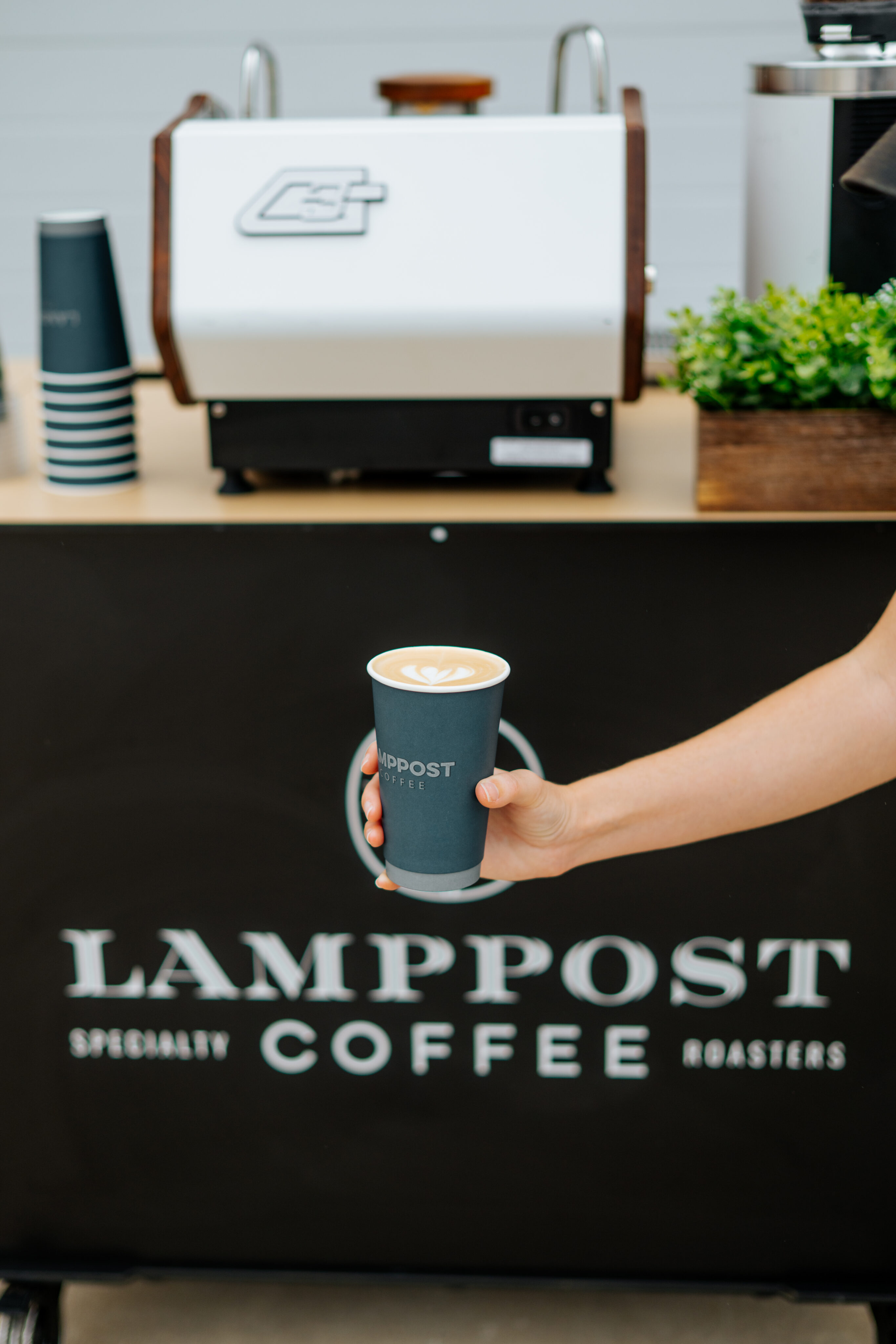 A hand holds a cup of coffee with latte art in front of a counter and espresso machine at Lamppost Coffee Roasters, where Office Services add convenience for busy professionals. - Lamppost Coffee
