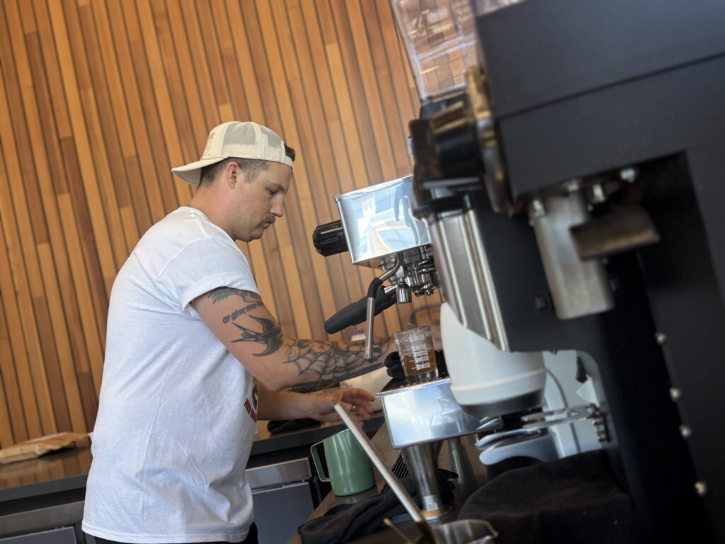A person wearing a backwards cap and white t-shirt uses a mobile coffee espresso machine in a coffee shop with wooden wall panels. - Lamppost Coffee
