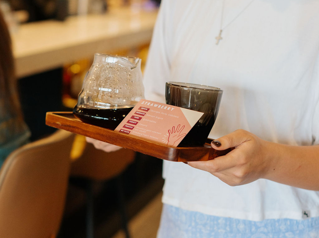 A person holds a wooden tray with a glass carafe, a black cup, and an information card labeled “STRAWBERRY” in Lamppost Coffee, highlighting the cafe’s specialty coffee offerings. - Lamppost Coffee