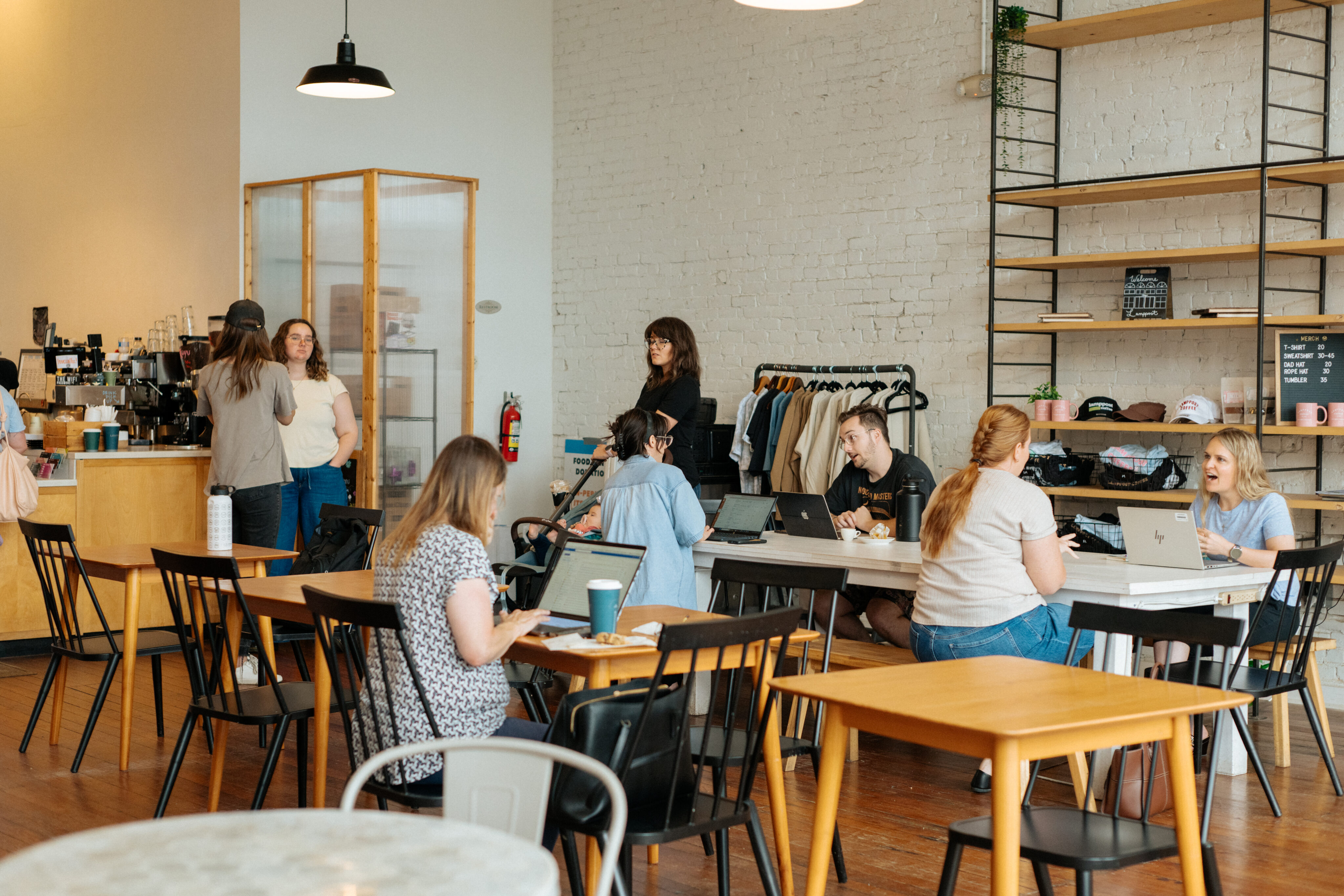 Several people are working, talking, and drinking coffee in a modern cafe with wooden tables, black chairs, and shelves on the wall. - Lamppost Coffee