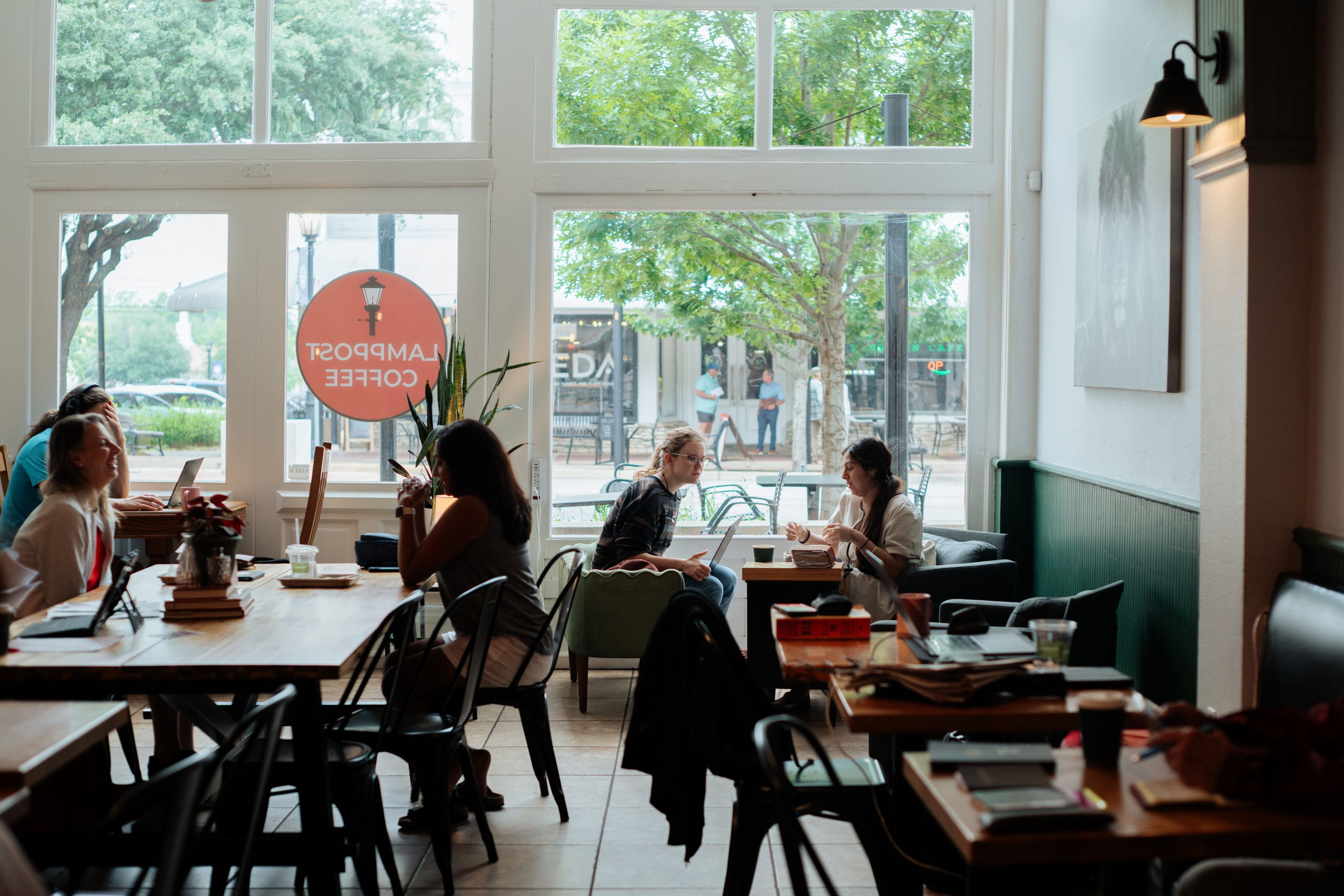 People sit at tables and work or talk in a bright, modern coffee shop with large windows and natural light in Downtown Round Rock. - Lamppost Coffee