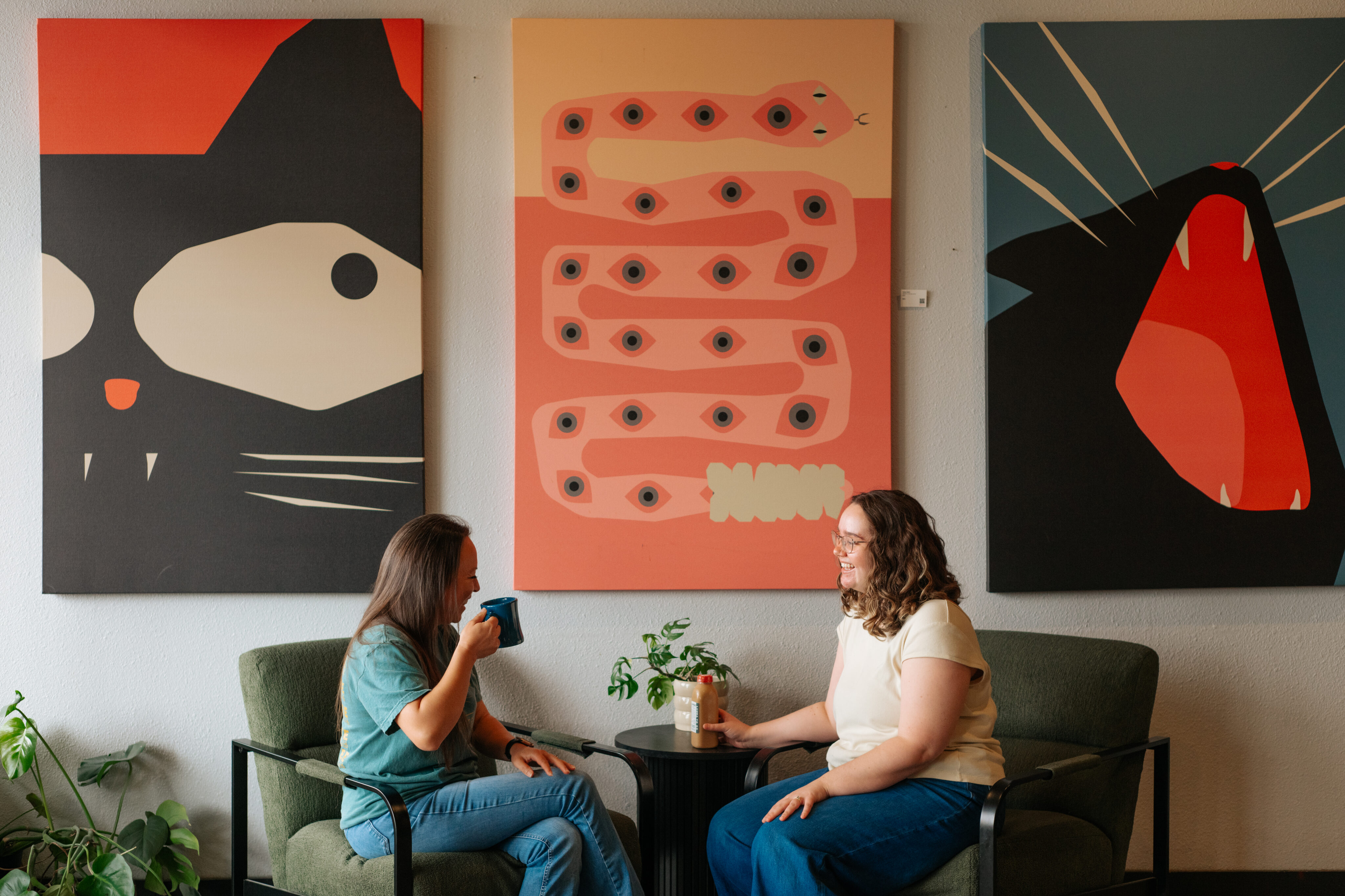 Two people sit in green chairs at a small table with drinks, in front of three large, colorful animal-themed artworks on the wall in a cozy Round Rock West setting. - Lamppost Coffee