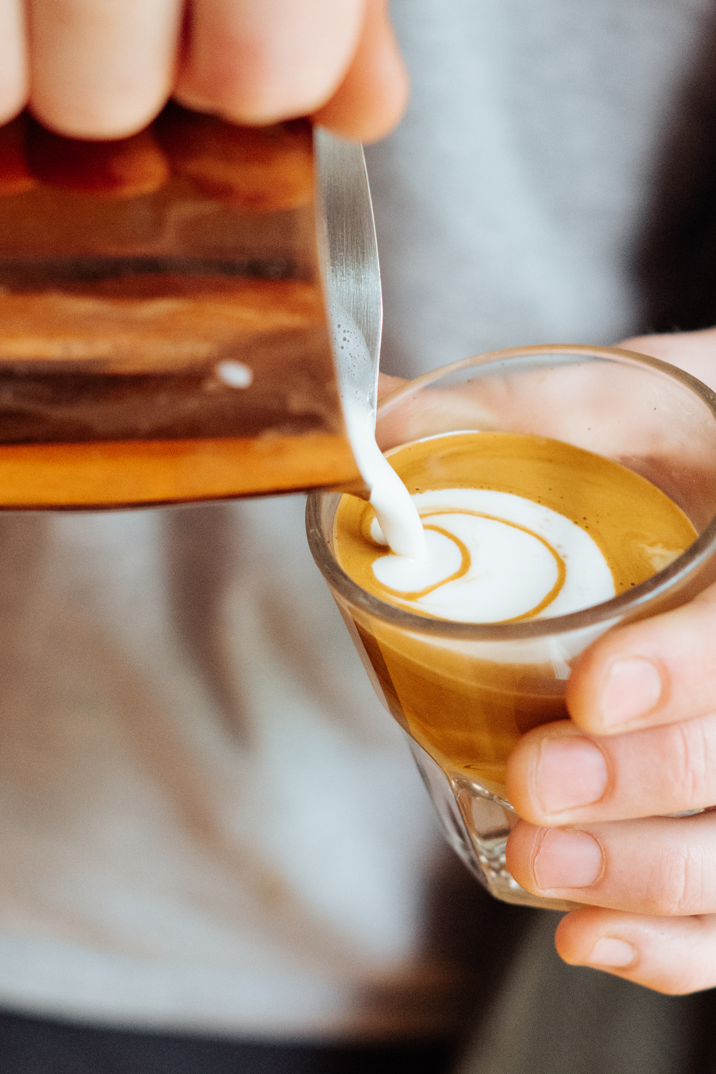 A hand pours steamed milk in a swirling pattern into a glass of espresso, creating latte art inside a bustling mobile cafe. - Lamppost Coffee