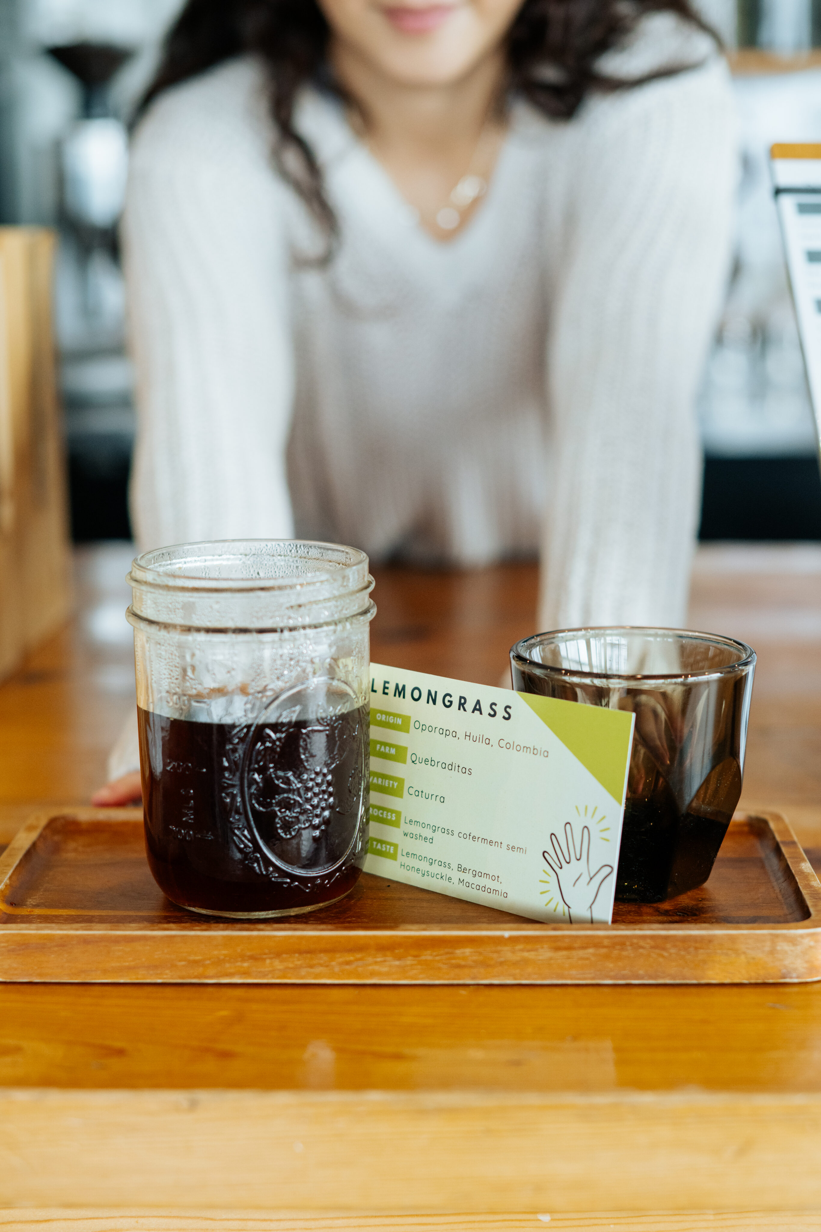 A person leans over a wooden counter with a glass jar of dark liquid, a small glass, and a card labeled "LEMONGRASS" on a tray. - Lamppost Coffee