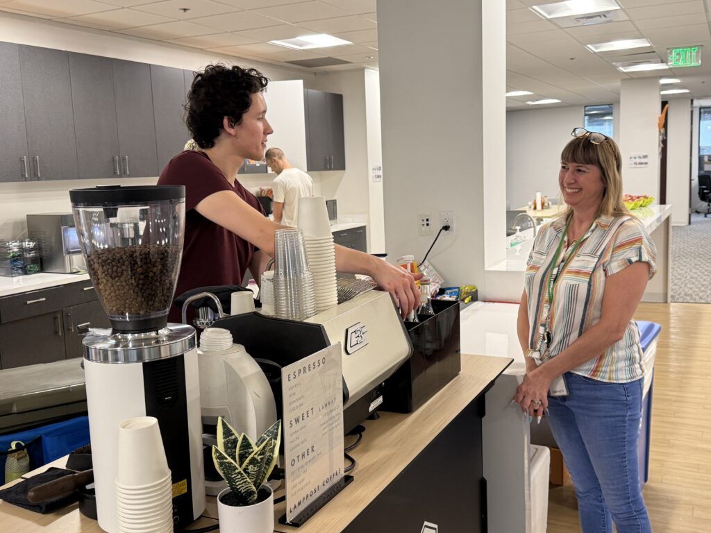A barista from a mobile coffee service prepares a coffee drink for a woman at a counter in an office kitchen, with another person in the background. - Lamppost Coffee