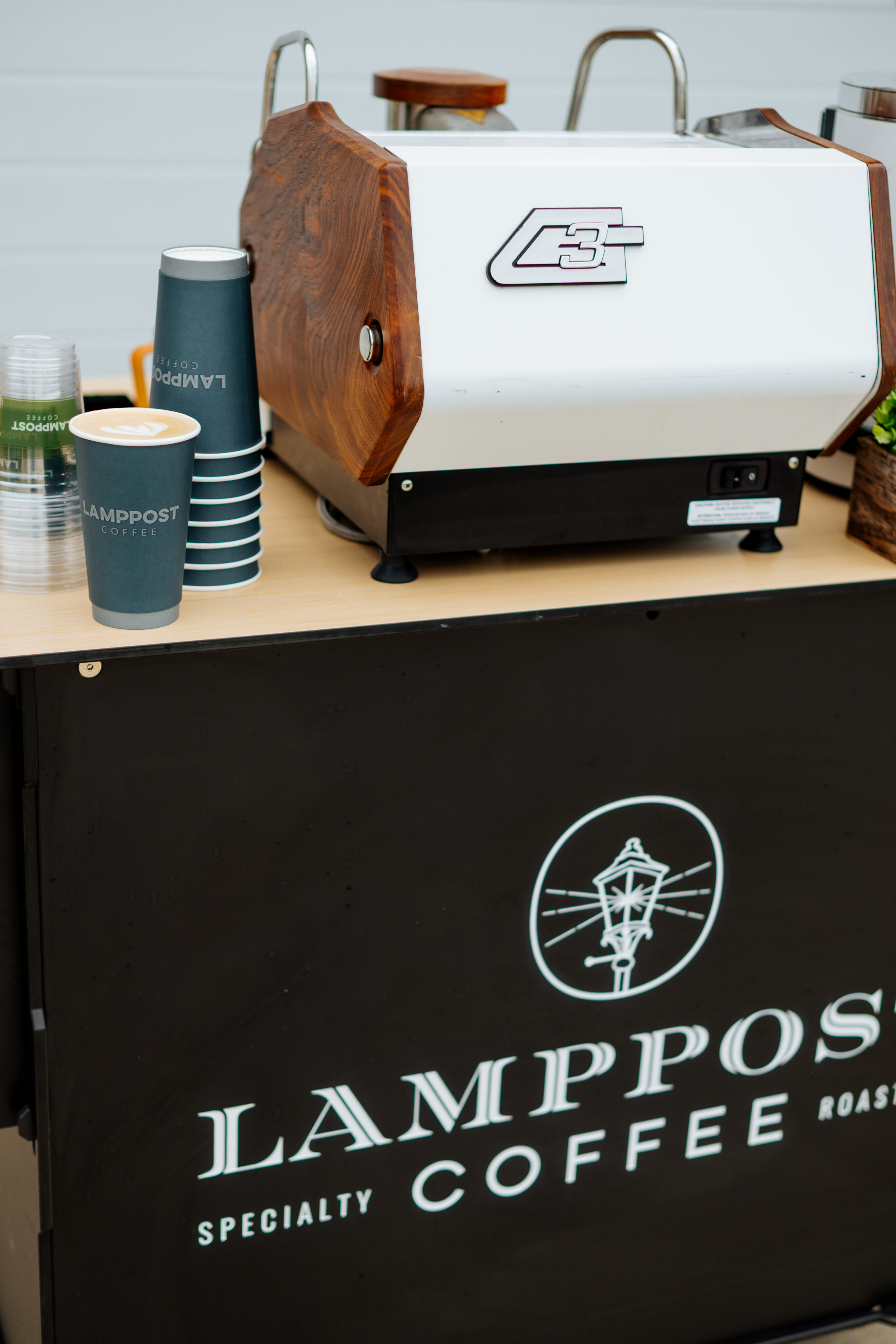 A coffee cart with Lamppost Coffee branding showcases specialty coffee, featuring a coffee machine, stacked paper cups, and lids on a wooden counter. - Lamppost Coffee