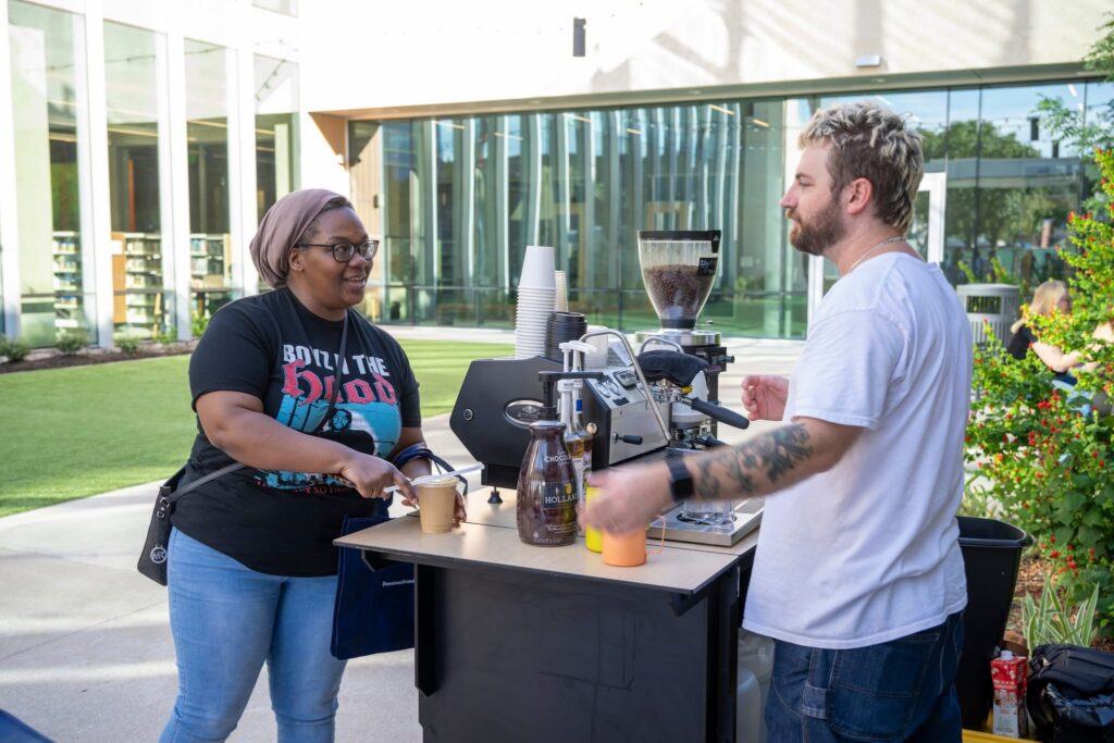 A woman receives a coffee from a barista at a mobile coffee truck, with a grassy courtyard and building in the background. - Lamppost Coffee