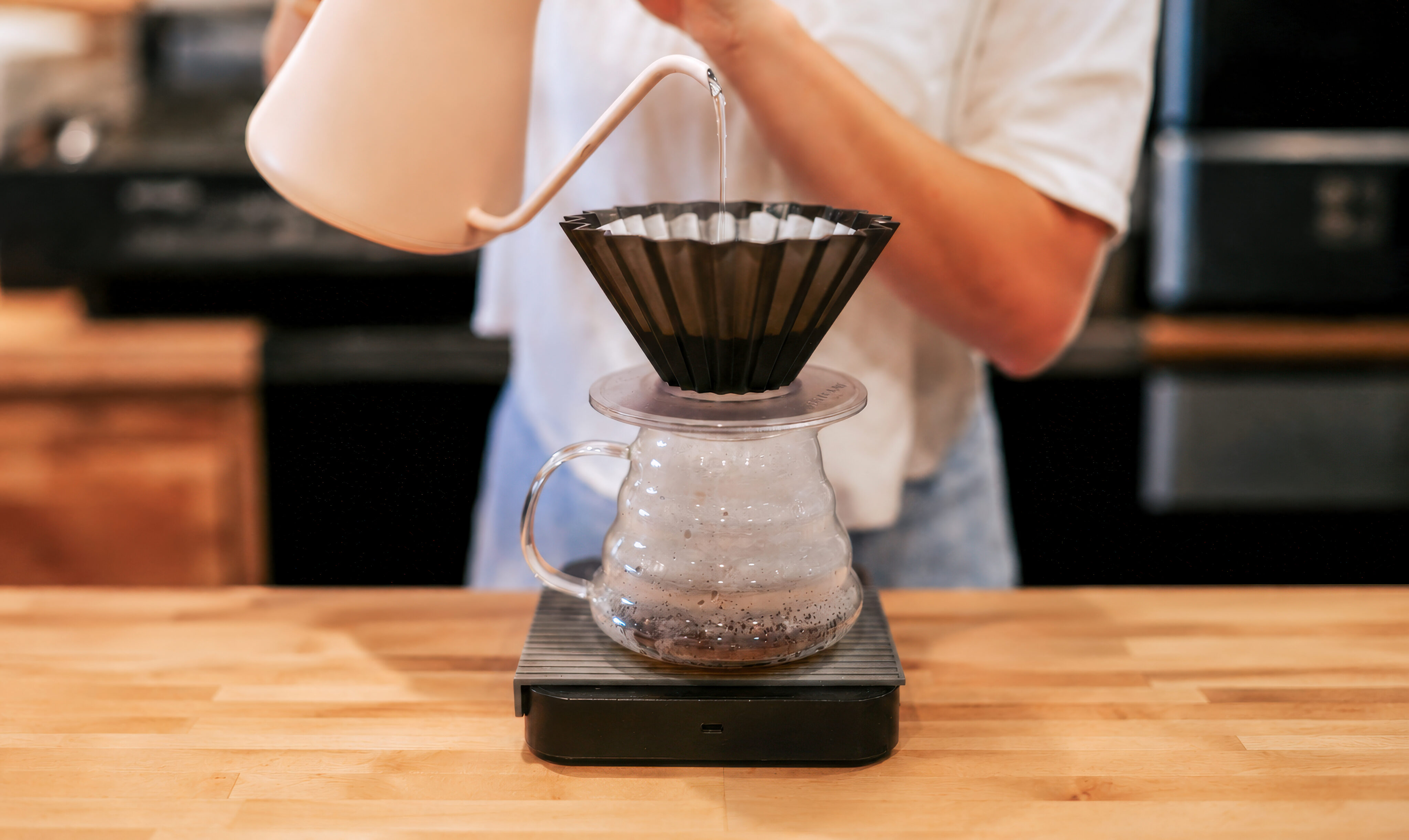 A barista at Lamppost Coffee pours hot water from a kettle into a dripper with a filter over a glass server on a scale, expertly preparing specialty coffee on a wooden counter. - Lamppost Coffee