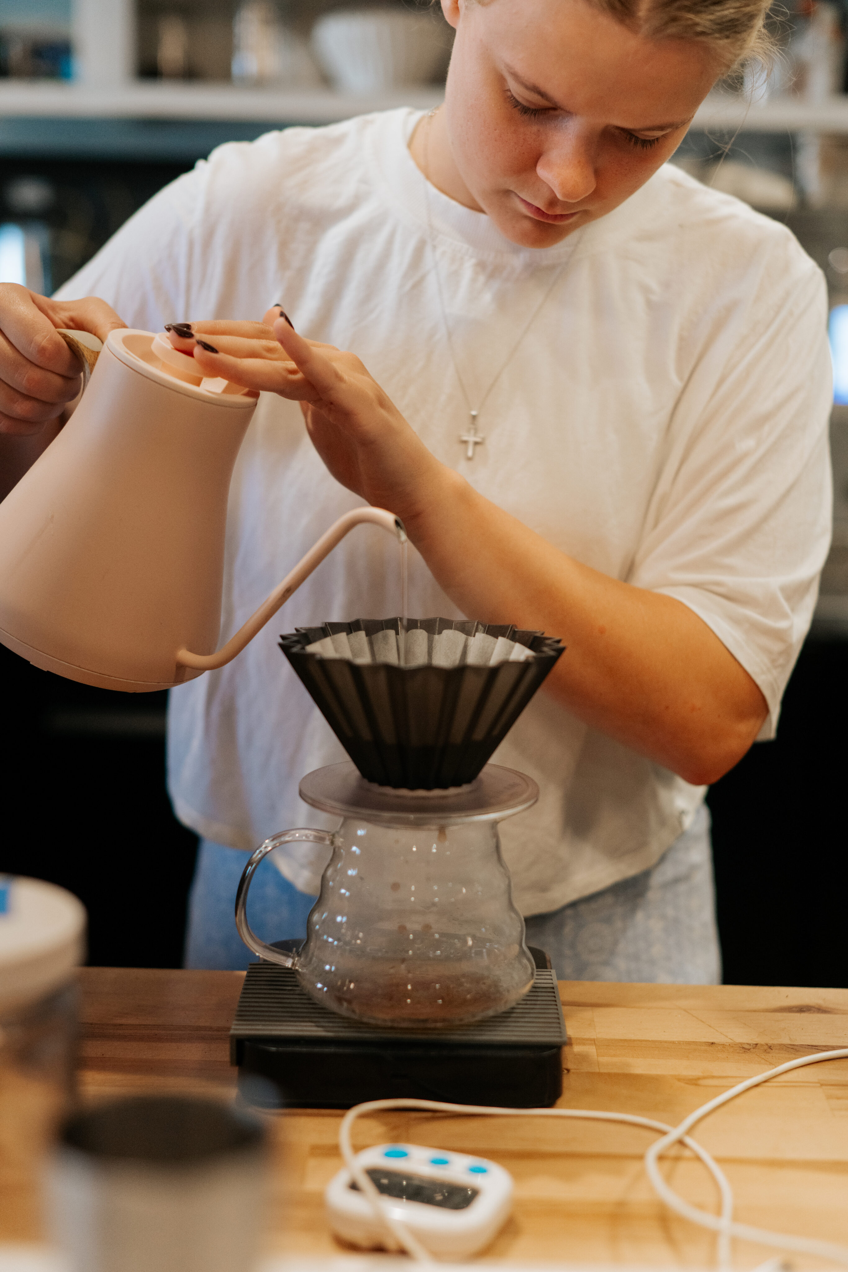 Person in a white shirt pours hot water from a kettle over a coffee dripper with a filter, making pour-over coffee on a scale—perfect for brewing that FAQ-worthy cup every time. - Lamppost Coffee