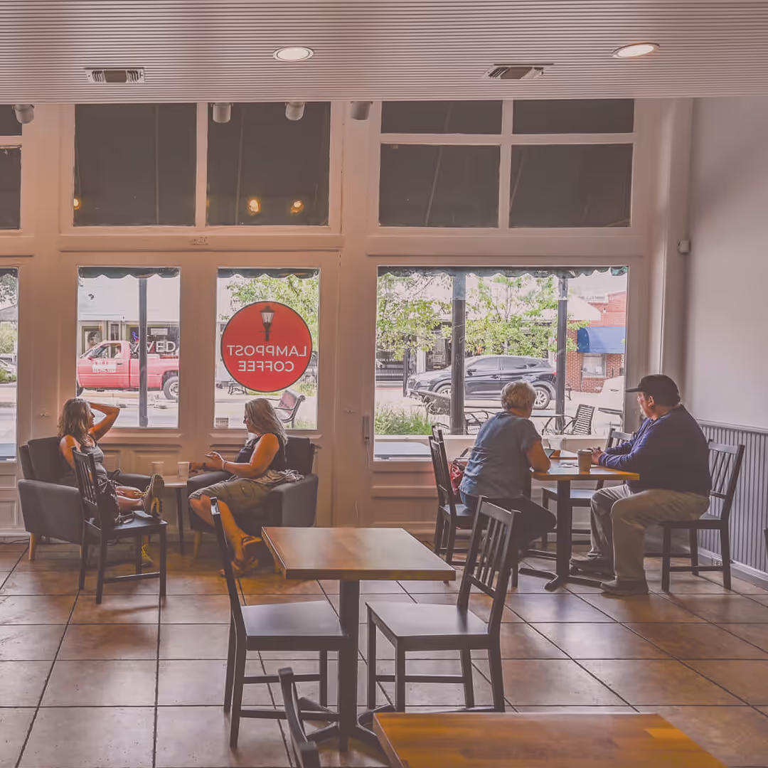 Two groups of people sit at separate tables inside a coffee shop near large windows, with a “Lamppost Coffee” sign visible—capturing a cozy moment in Downtown Round Rock. - Lamppost Coffee