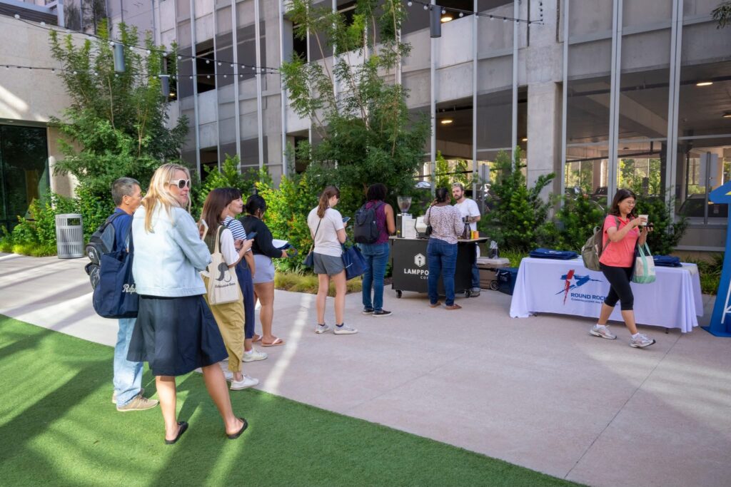 People stand in line outdoors near a table with brochures and a man serving coffee from a mobile coffee setup; a building and greenery are in the background. - Lamppost Coffee