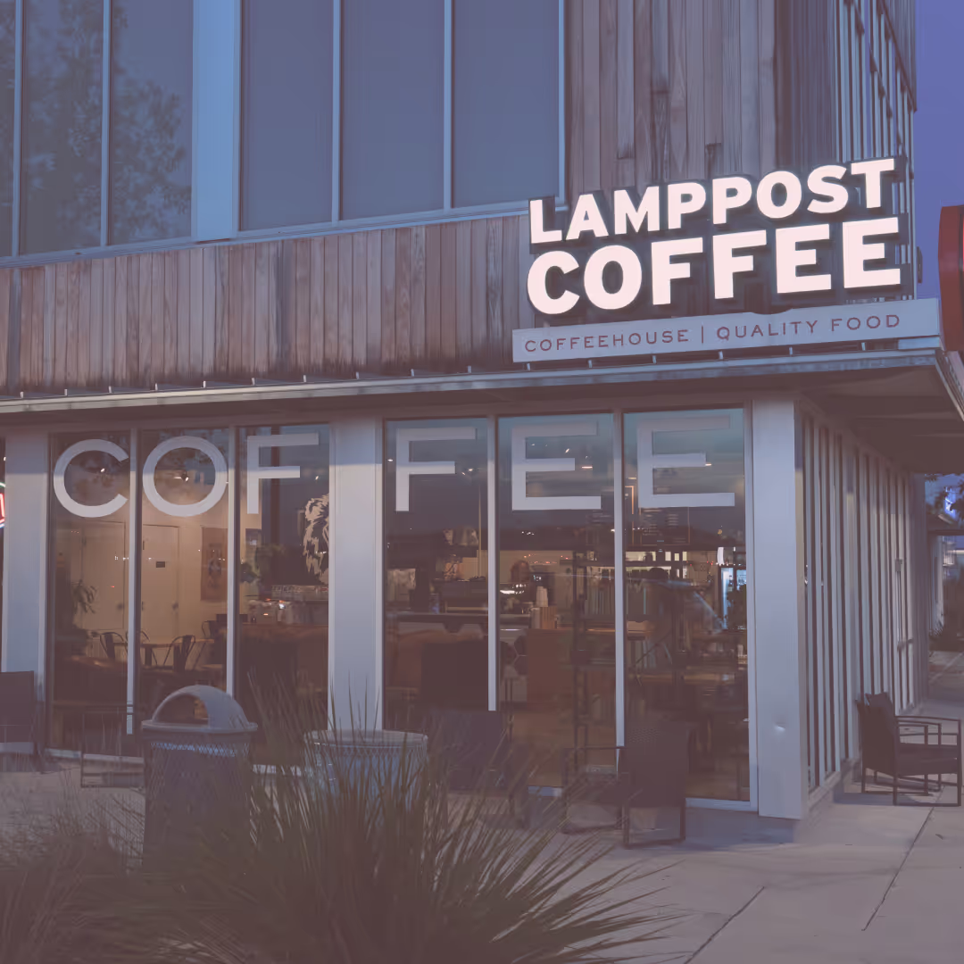 Street view of Lamppost Coffee, a modern coffeehouse with large windows, wood siding, and seating visible inside. Sign reads "Lamppost Coffee: Coffeehouse | Quality Food. - Lamppost Coffee